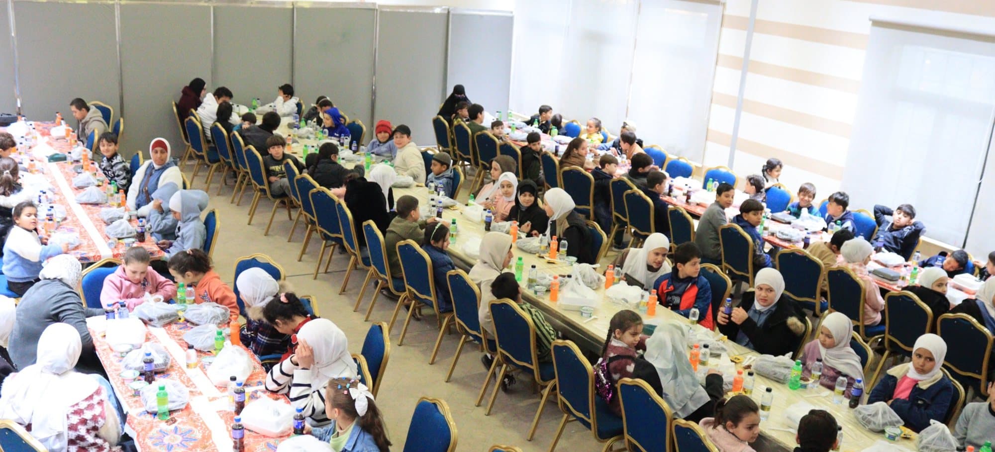 Children and volunteers seated at long Iftar tables in a banquet hall in Ma'an